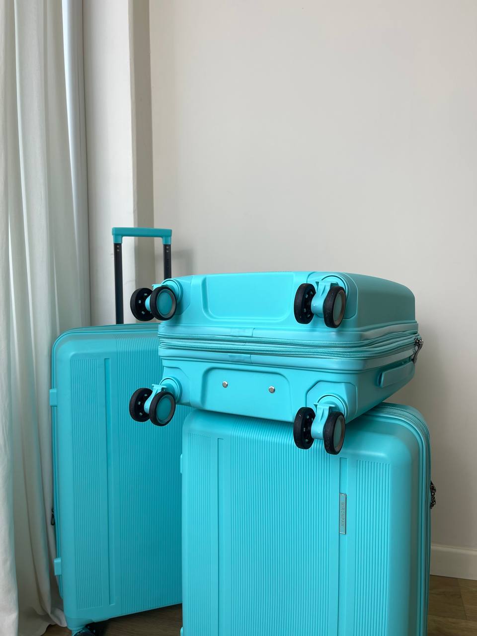 Три бирюзовых чемодана с жёстким корпусом Mironpan Classic, два стоят вертикально, один лежит сверху, на фоне белой стены и занавески.